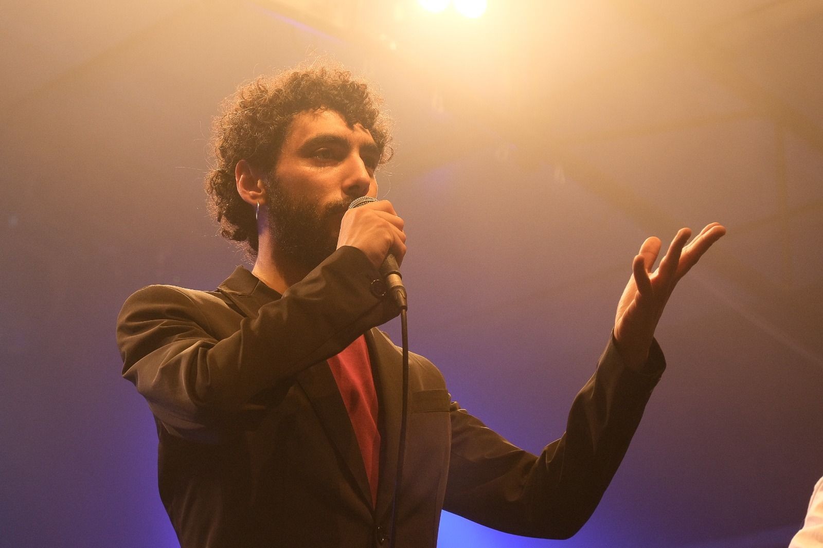 Person in a suit holding a microphone, speaking on stage with warm lighting and a spotlight above.
